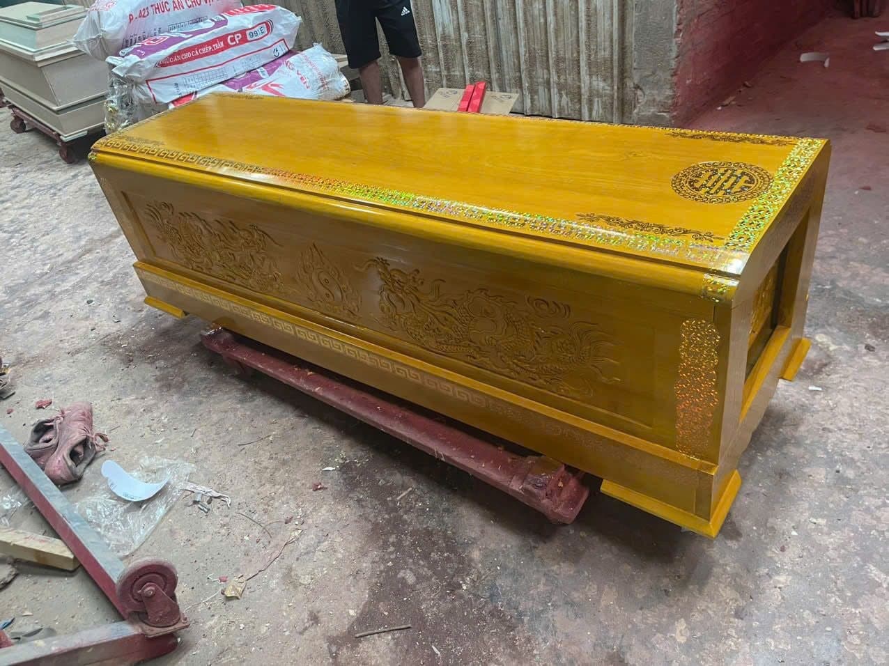 Quan tài gỗ vàng tâm sơn cánh gián chạm lưỡng long, đặt trên xe đẩy trong xưởng mộc